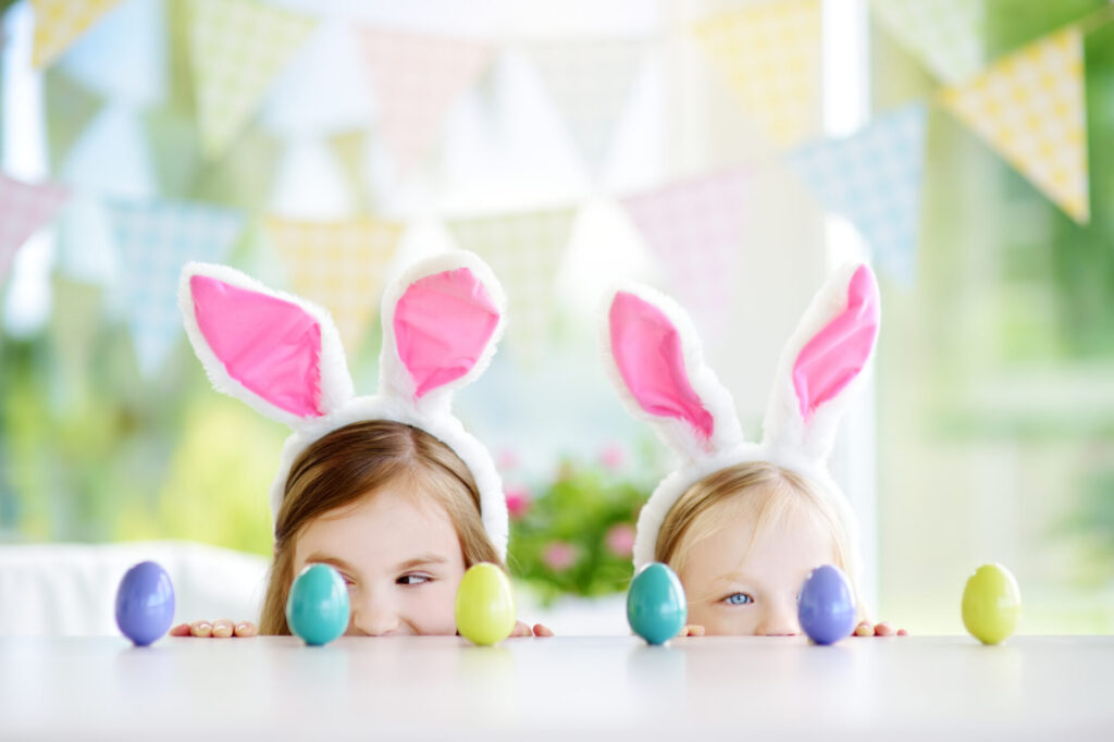 Two cute little sisters wearing bunny ears playing egg hunt on Easter. Adorable children celebrate Easter at home.