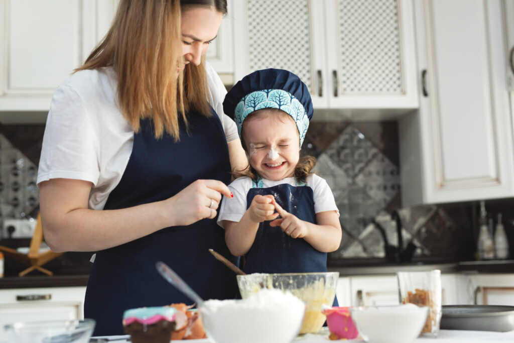 Cute little girl and her beautiful mom in matching aprons and caps play and laugh while kneading dough in the kitchen.