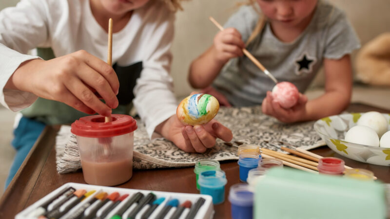 Close up of hands of adorable little boy painting colorful Easter eggs together wih his sister while spending time at home. Family, creativity, leisure activity concept. Horizontal shot