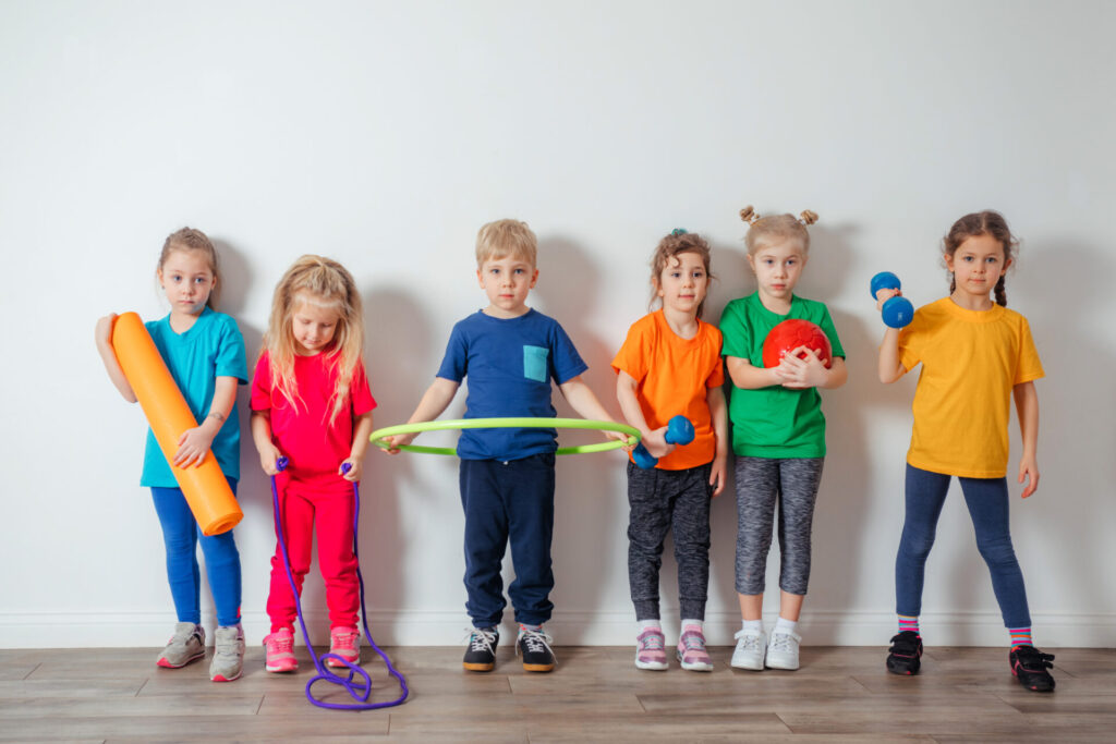 Group of cheerful young kids in colorful and comfortable sportswear posing next to white wall each holding one item of sport equipment. Active kids look tired after training.